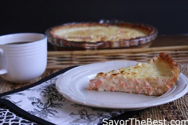 A slice of salmon quiche on a white plate next to a cup of coffee. 
