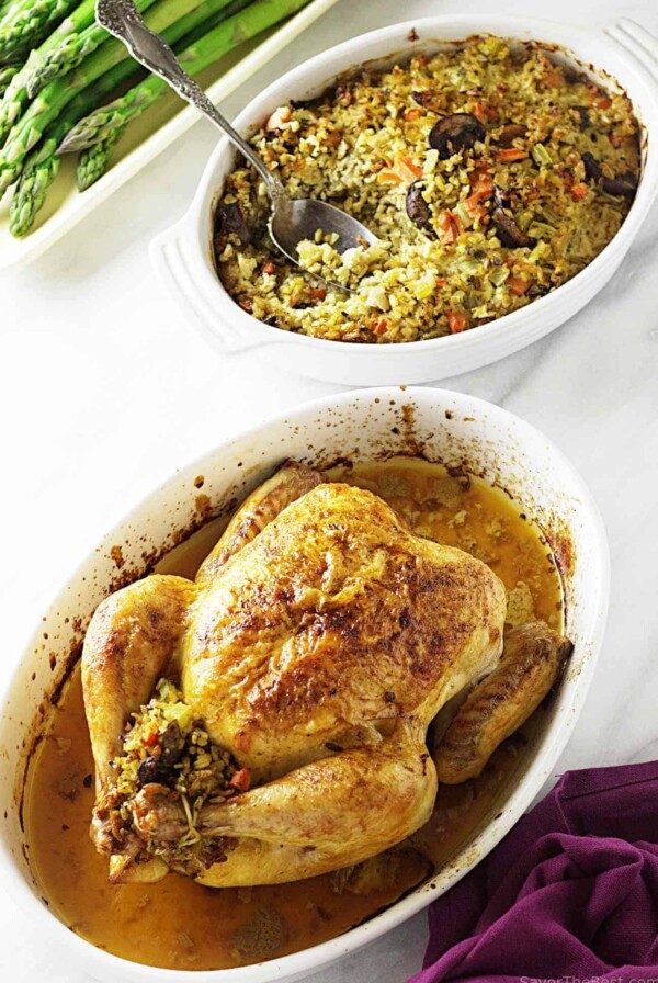 A roasted chicken in a casserole dish next to a dish of freekeh stuffing.