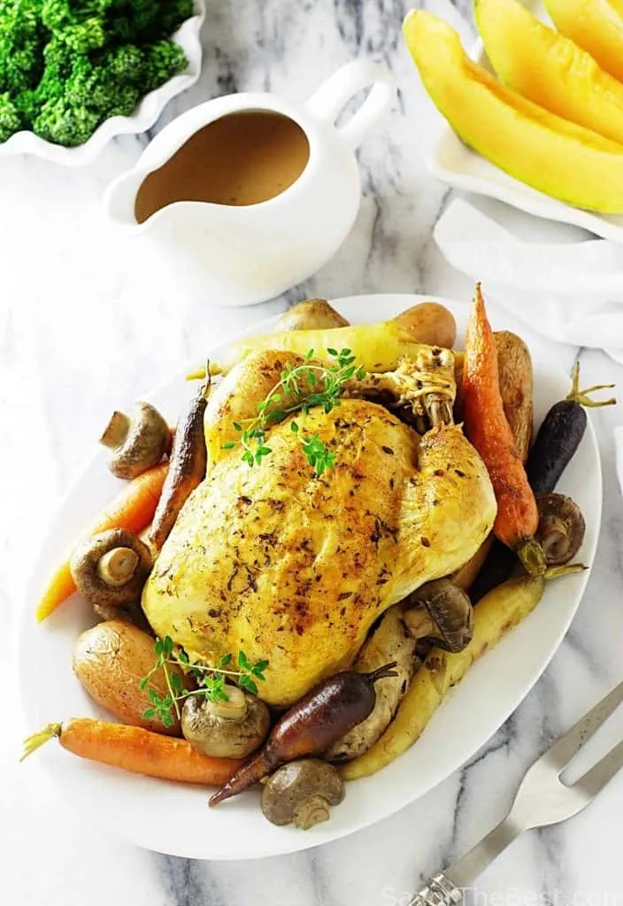 A whole roasted chicken on a plate with vegetables next to a pitcher of gravy. 