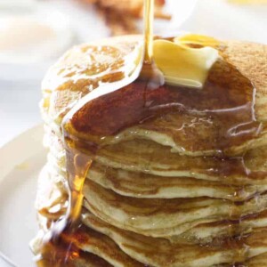 syrup pouring on a stack of pancakes with bacon in the background