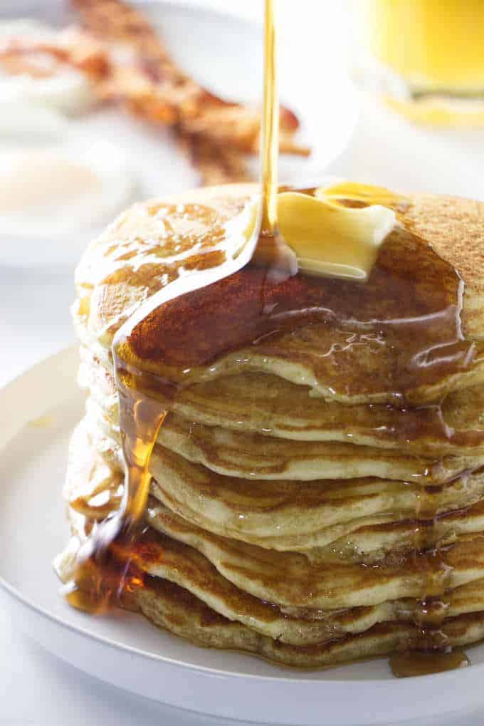 syrup pouring on a stack of pancakes with bacon in the background