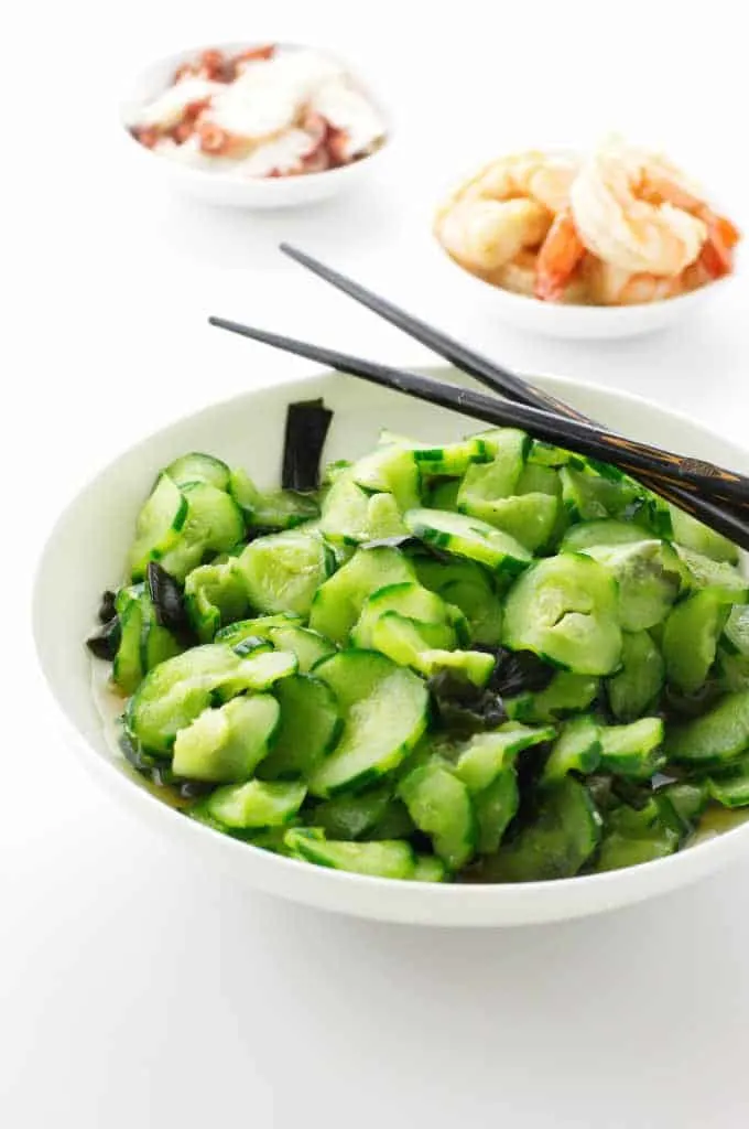 A bowl of cucumber salad and chopsticks. Cooked octopus and shrimp in dishes in background