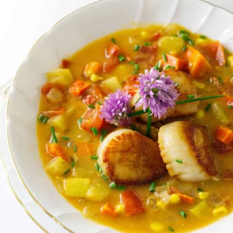 A bowl of vegetable chowder and three scallops, garnished with fresh chive blooms