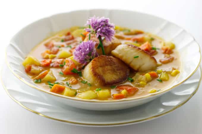 a bowl of vegetable chowder with 3 scallops and garnished with fresh chive blooms