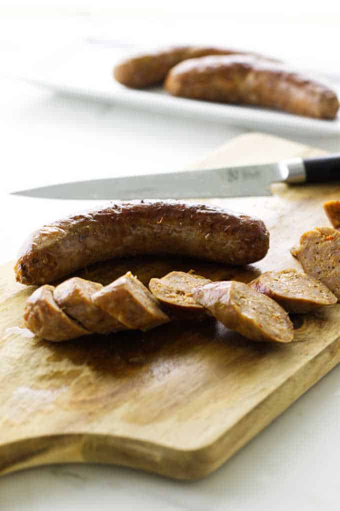 Sliced and whole andouille sausage on cutting board with knife. 2 sausages on plate in the background.