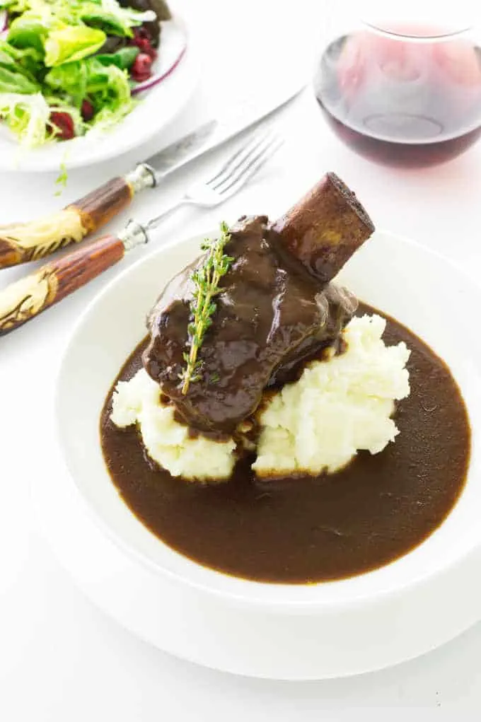 Dish with sauce, mashed potatoes, short ribs, wine and salad in background