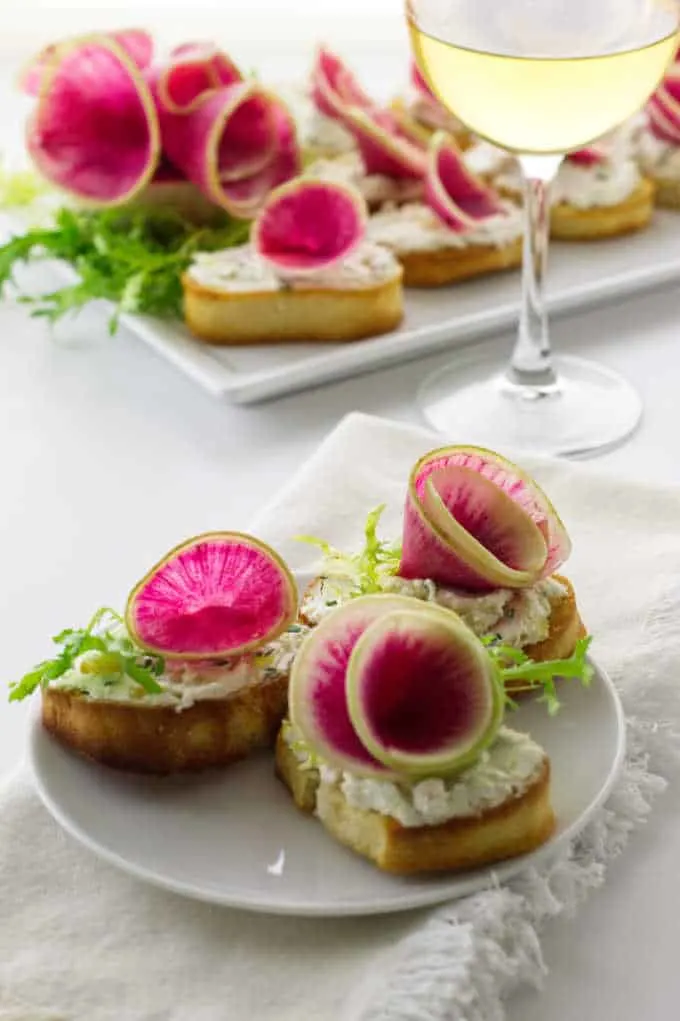 three watermelon radish and goat cheese crostini servings on plate with a plate in background and glass of wine