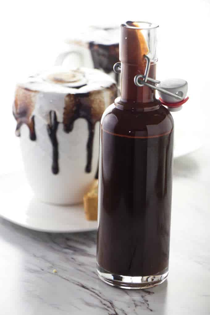 A bottle of homemade chocolate syrup with a cup of hot chocolate in the background.