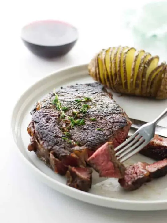 Ribeye steak with fork holding a bite, hasselback potato and glass of wine in background