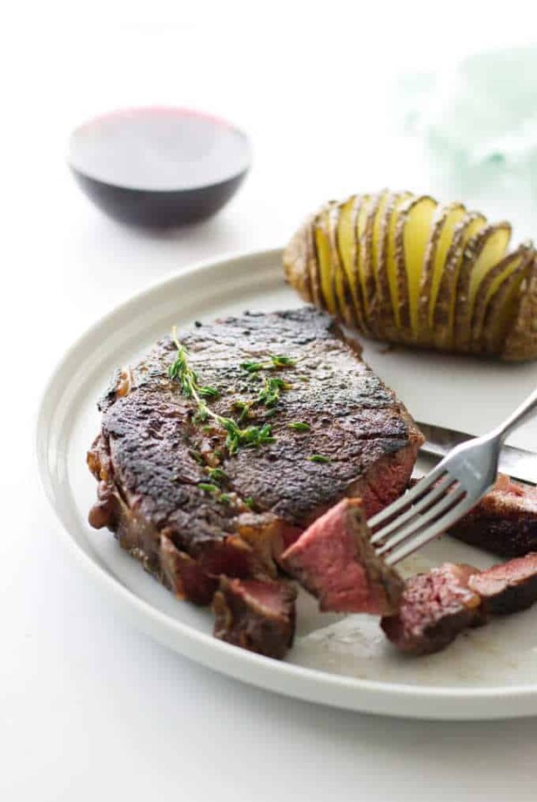 Ribeye steak with fork holding a bite, hasselback potato and glass of wine in background