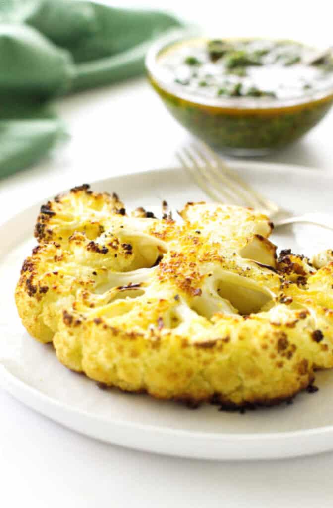 Thick roasted cauliflower steak on plate with fork. Dish of chimichurri sauce and napkin in background.