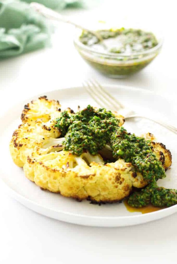 Plate with cauliflower steak and chimichurri sauce, fork and a dish of sauce in background