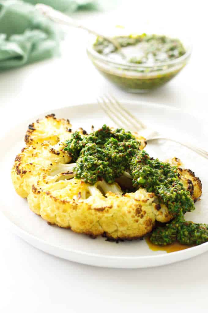 Plate with cauliflower steak and chimichurri sauce, fork and a dish of sauce in background