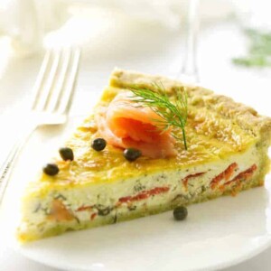 Serving of quiche on a plate with fork, napkin and wine in background