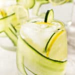 A glass of cucumber lemon water with a pitcher in the background.