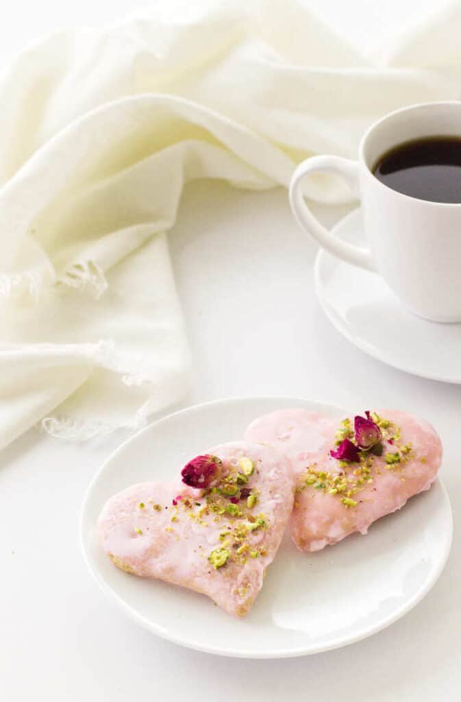 Overhead view of two pink heart-shaped cookies, decorated with dried rosebuds and chopped pistachios