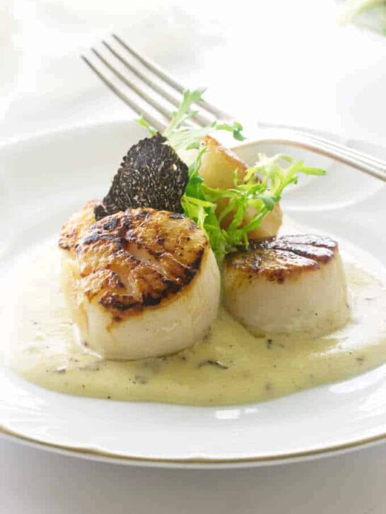 A plate of seared scallops with a black truffle slice and black truffle sauce.
