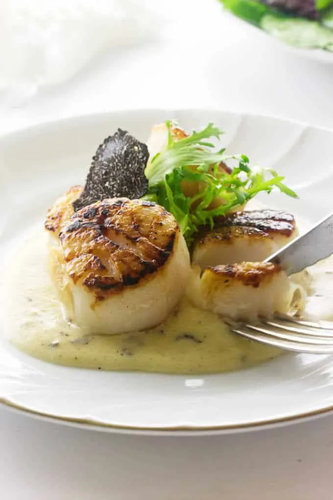 Slicing a seared scallop and dipping it in black truffle sauce garnished with frisée greens and a slice of truffle