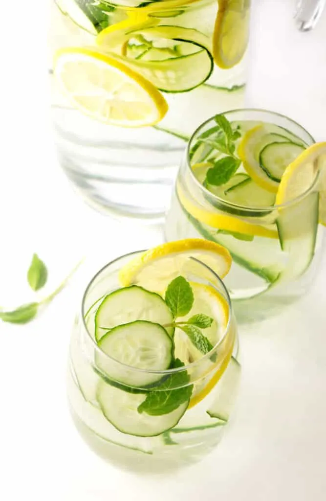 Two glasses of cucumber lemon water with a pitcher in the background.