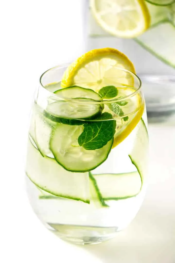 A glass of cucumber lemon water with a pitcher in the background.