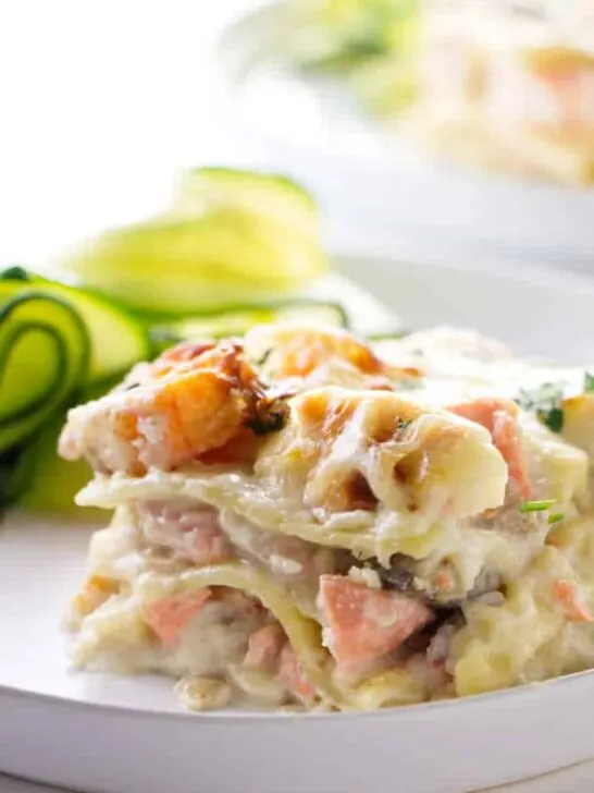 A slice of seafood lasagna with zucchini in the background.