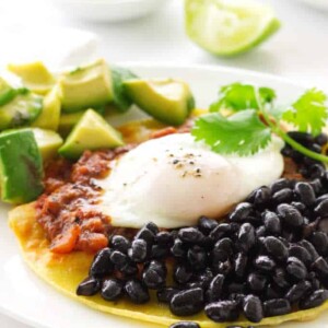 Corn tortilla with black beans, salsa, egg and avocado chunks. Lime wedges and corn tortillas in background.