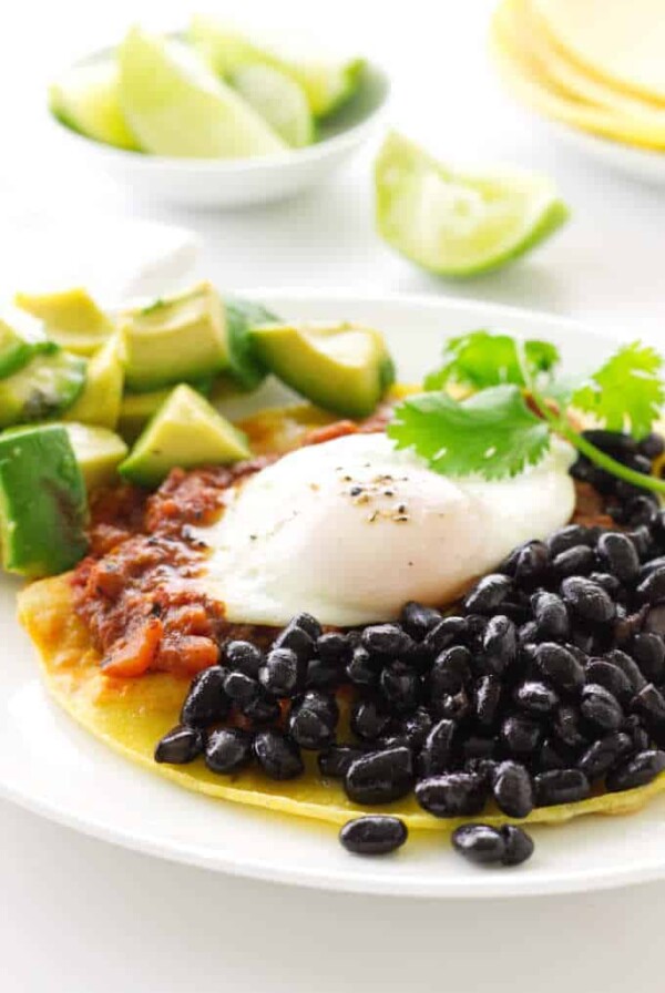 Corn tortilla with black beans, salsa, egg and avocado chunks. Lime wedges and corn tortillas in background.