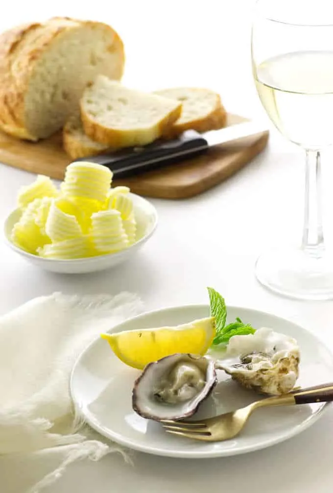 2 oysters on appetizer plate with fork, lemon wedge and mint. Bread, butter curls and glass of wine in background