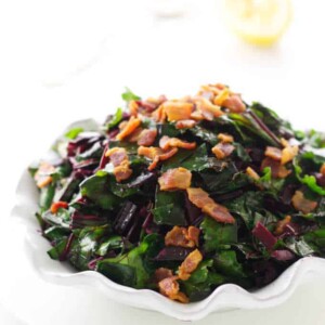 Close up view of side dish of beet greens with bacon bits. Spoon and lemon in background