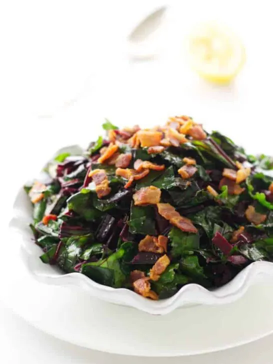 Close up view of side dish of beet greens with bacon bits. Spoon and lemon in background