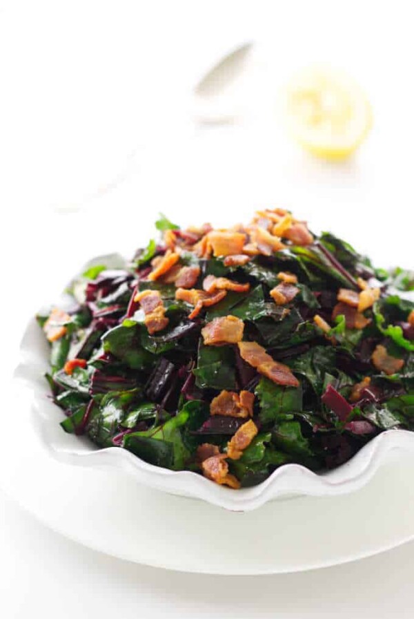 Close up view of side dish of beet greens with bacon bits. Spoon and lemon in background