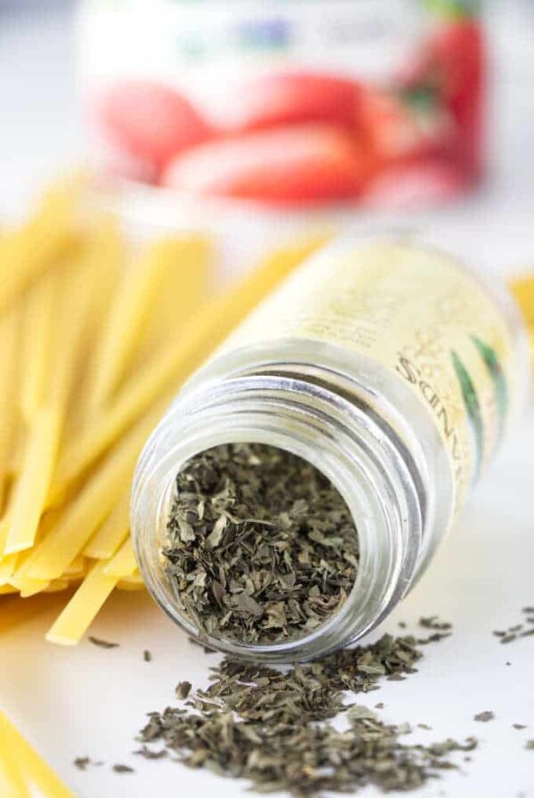 Dry basil and dried pasta with a can of tomatoes in the background.