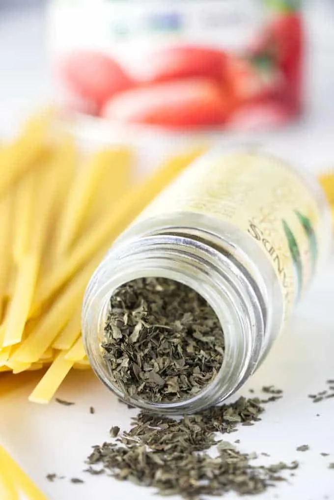 Dry basil and dried pasta with a can of tomatoes in the background.