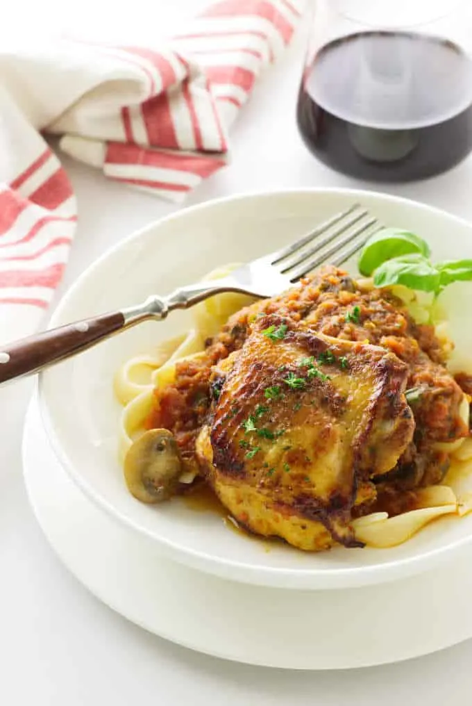 Overhead view of a serving of chicken thigh in Italian tomato sauce with a fork. Napkin and a glass of red wine in background