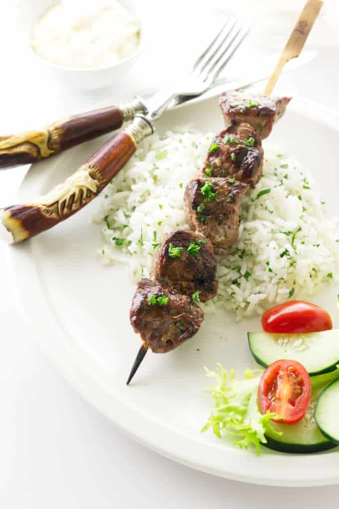 overhead view serving of grilled lamb kabobs, rice, salad, knife/fork and garlic aioli sauce in background