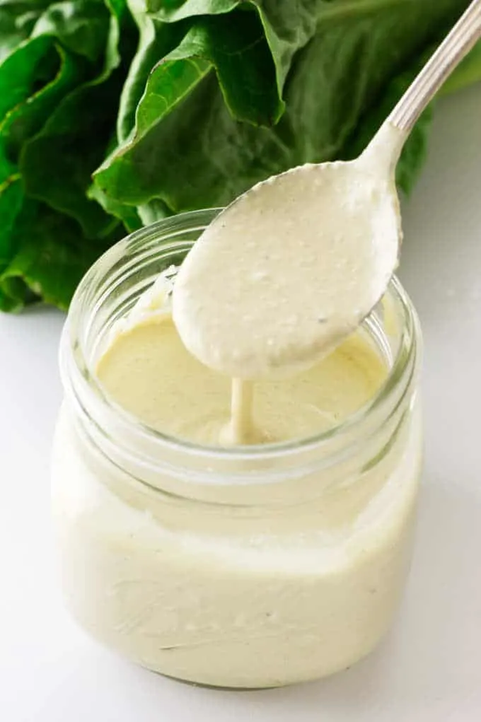 Jar of Caesar Salad Dressing with a spoonful, romaine lettuce in background