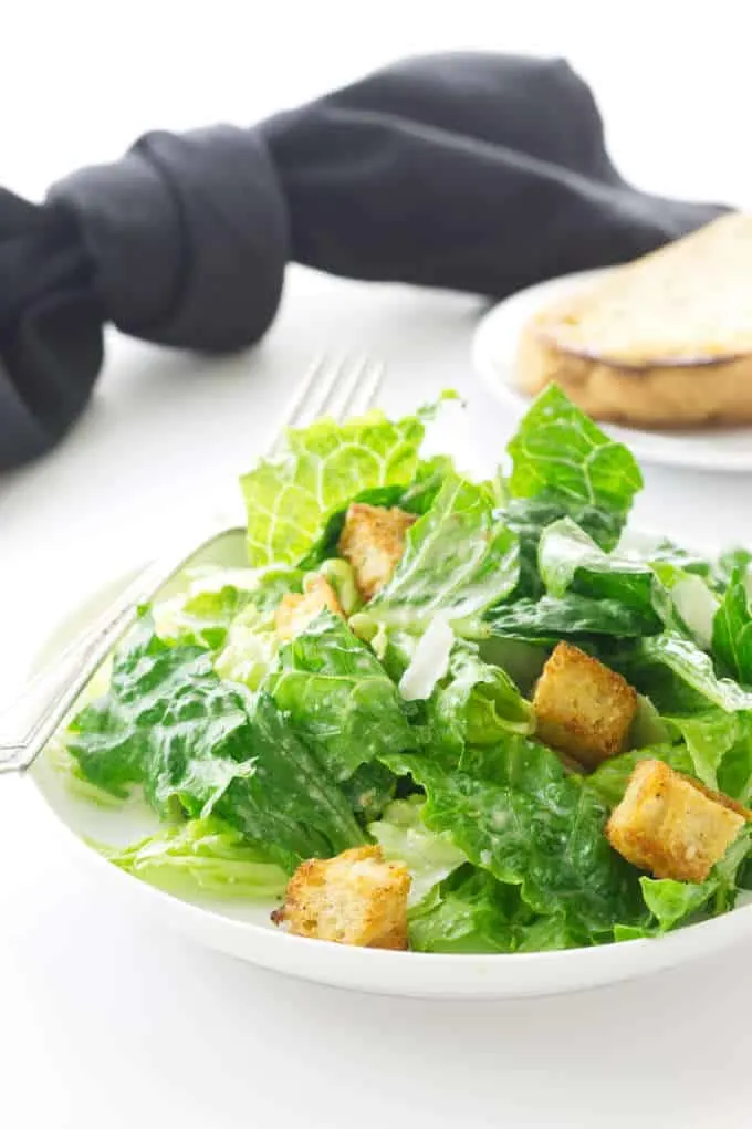 Serving of Caesar Salad on a plate with fork. Saucer with bread in background with black napkin h