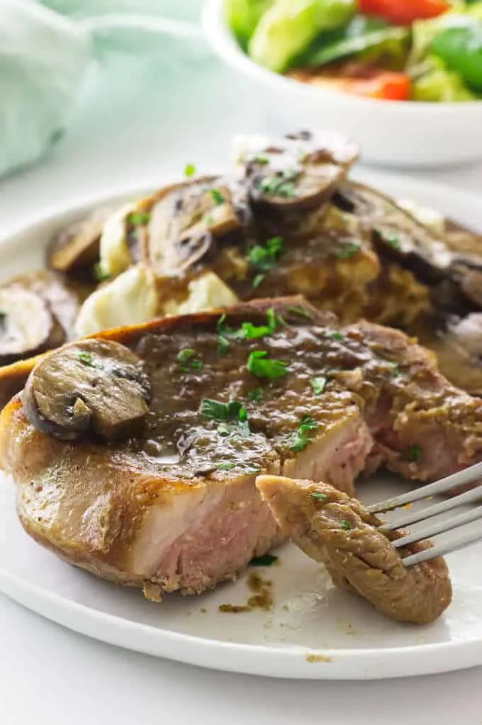 A pork chop serving on plate with mushrooms and mashed potatoes, a uicy bite of pork chop on fork, salad and napkin in background.