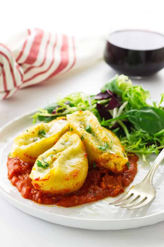 a serving of cheese stuffed pasta shells with marinara sauce, side salad and a fork. Napkin and glass of wine in background.