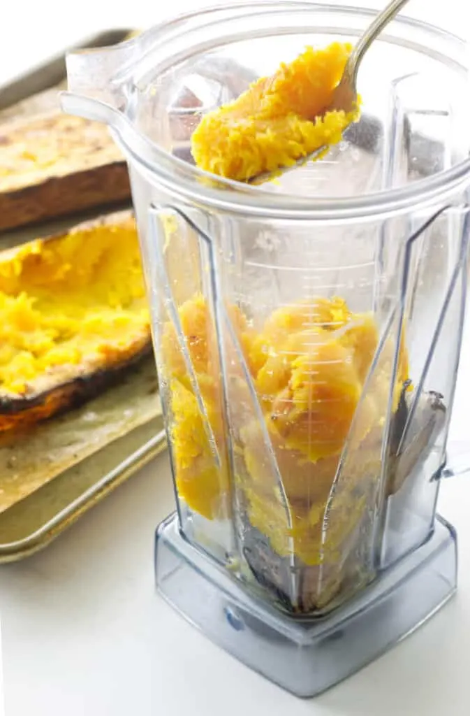Blender with roasted butternut squash, spoon with squash. Tray of baked squash in background