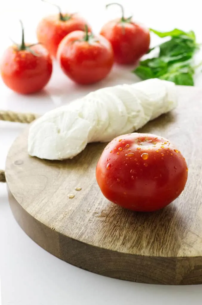 Cutting board with tomato and sliced mozzarella, tomatoes and basil in background