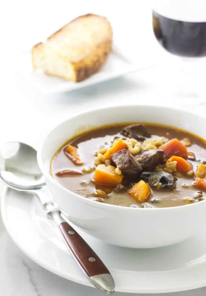 A serving of beef barley soup with a glass of wine and a slice of bread.