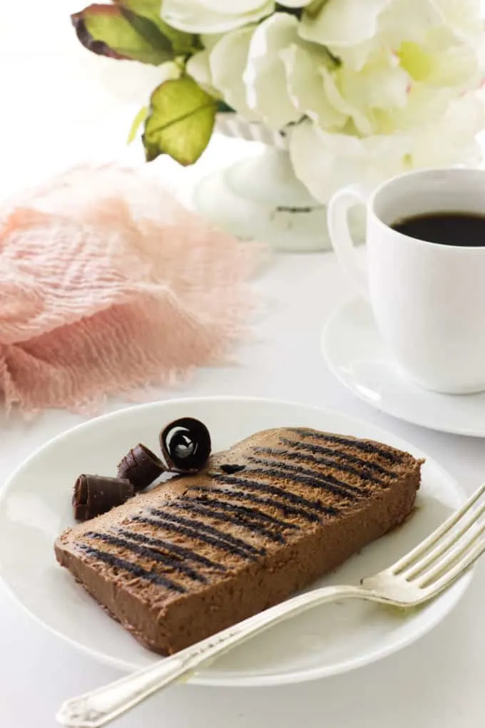 Close up view of a slice of double chocolate icebox cake on a dessert plate w/fork, cup of coffee, napkin and flowers in background