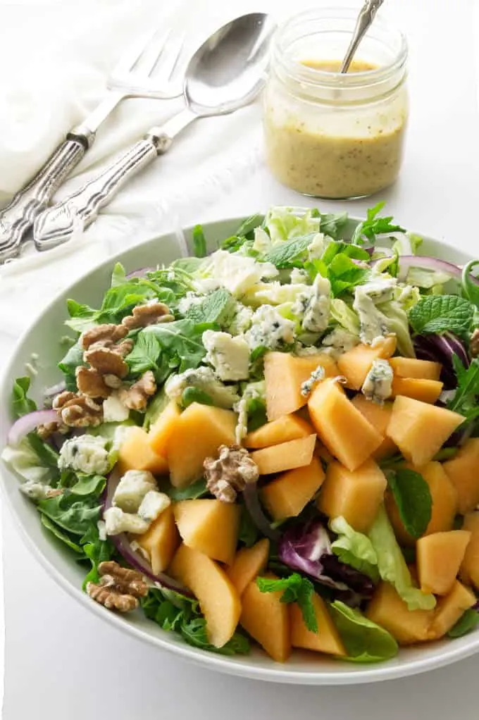 A large green salad with cantaloupe and blue cheese and a jar of dressing in the background.