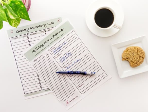 A holiday menu planner on a desk with coffee and a cookie and a pen.