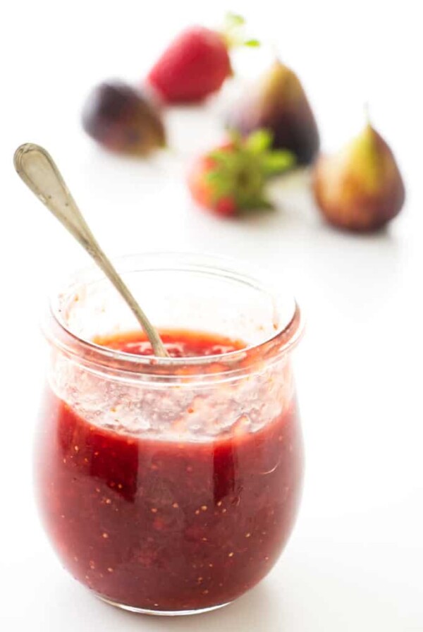 A jar of strawberry fig jam with fresh strawberries and figs in the background.