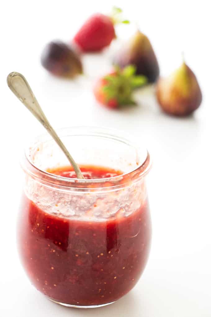 A jar of strawberry fig jam with fresh strawberries and figs in the background.