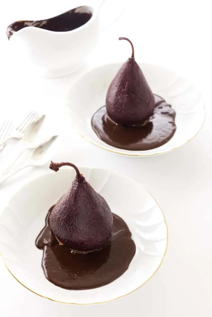 Two red wine poached pears in individual serving plates with chocolate sauce