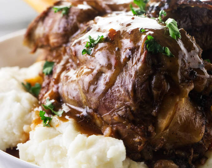 A lamb shank on a bed of mashed potatoes with gravy on top.