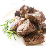 View of pan fried lamb chops on a plate with fresh rosemary garnish. Serving fork in background